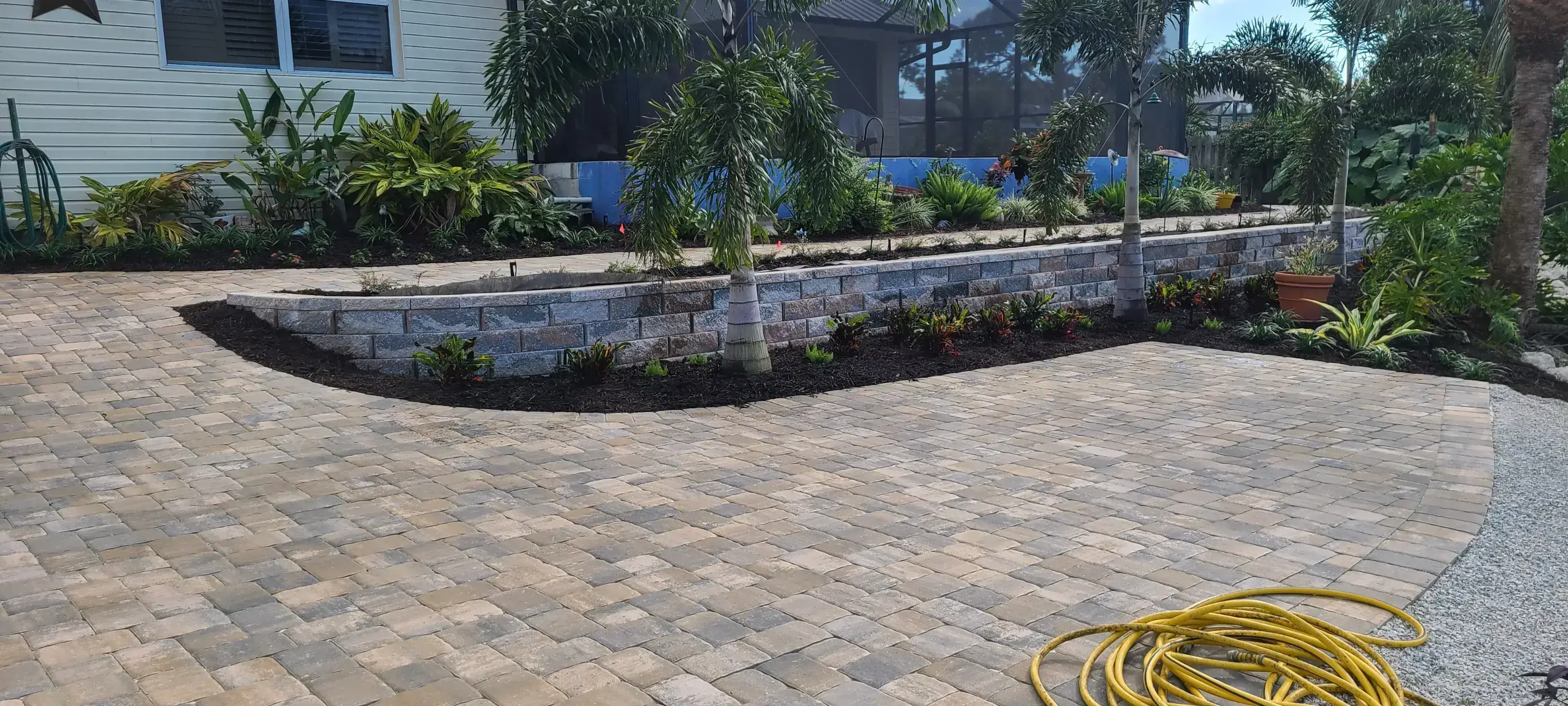 Palm tree and flowering plants along a stone retaining wall beside a paved driveway.