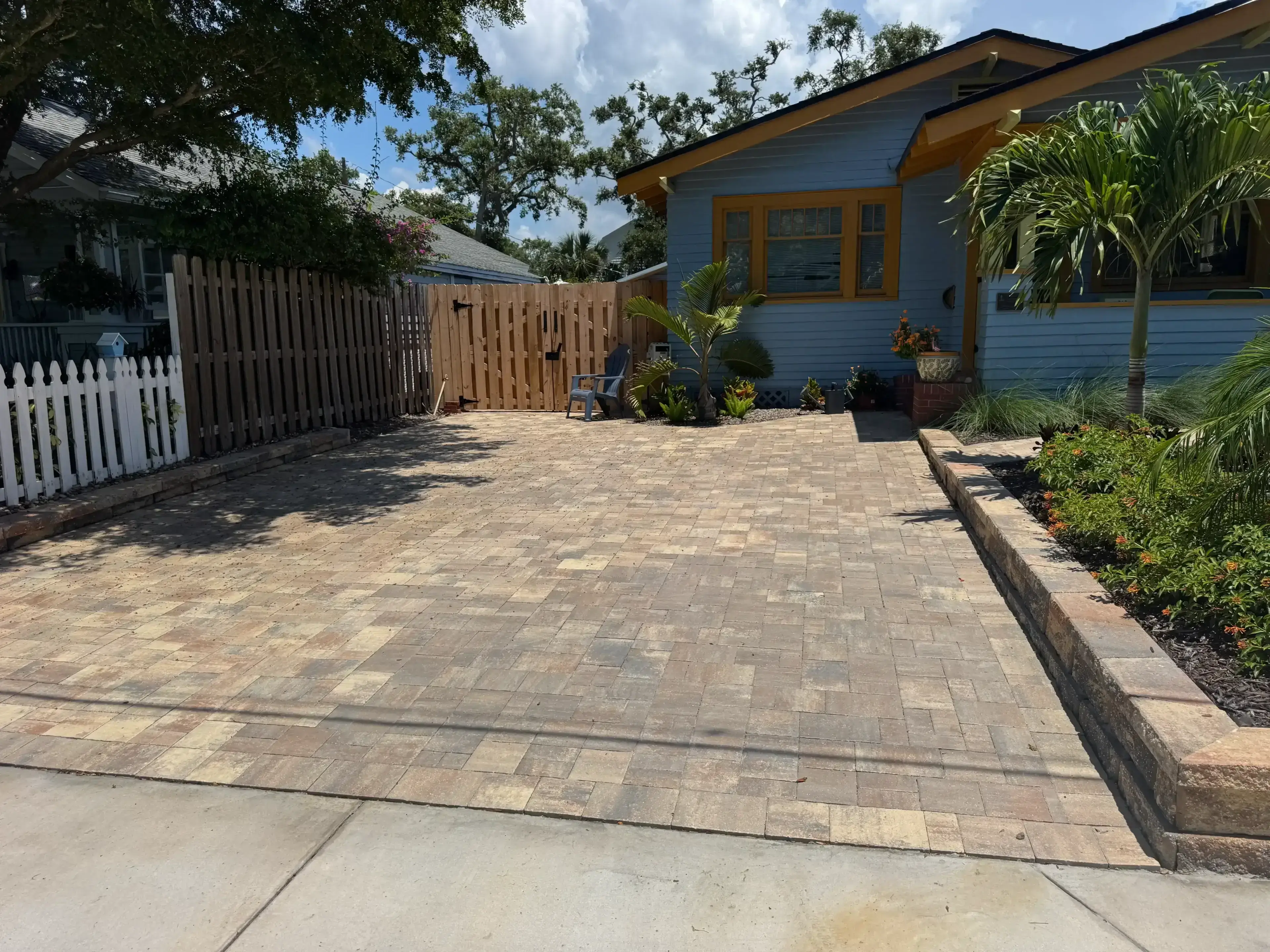 A paved driveway in front of a yellow and blue house with plants around.
